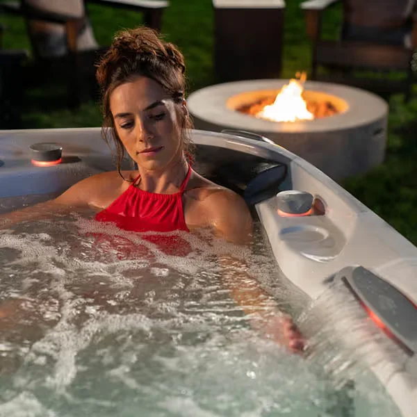 A woman enjoying a quiet evening soak in a Sundance hot tub beside a fire pit